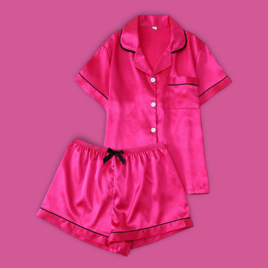 A two-piece women's pajama set in bright pink satin, featuring a short-sleeve button-front top with black piping and matching shorts with a black drawstring and black piping, displayed on a solid pink background.